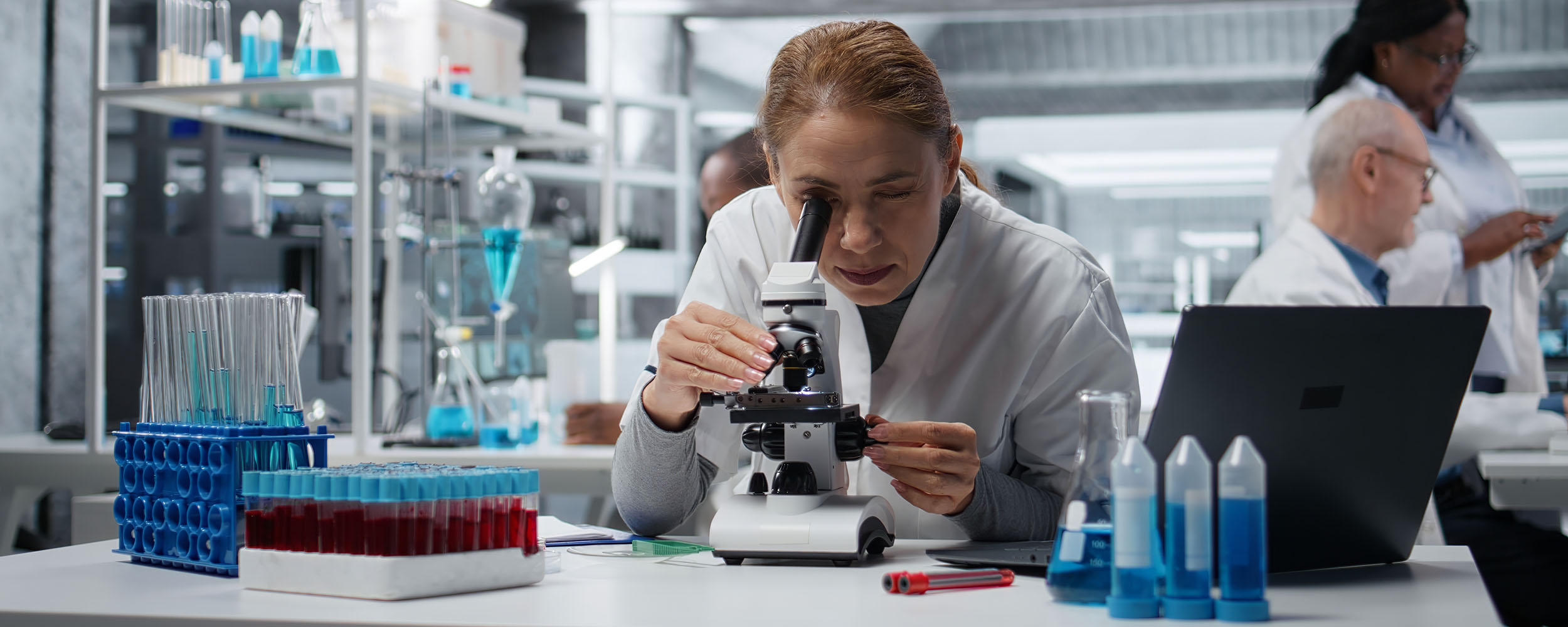 Molecular biologist in medical laboratory using scientific microscope for cellular analysis. Female researcher doing sample examination inside lab with optical device gear, camera A